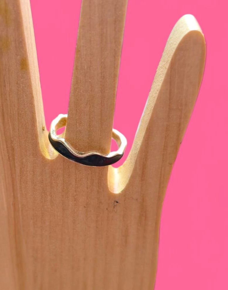 Anillo con diseño de ola negra sobre un soporte de madera con forma de mano, sobre un fondo rosa.
