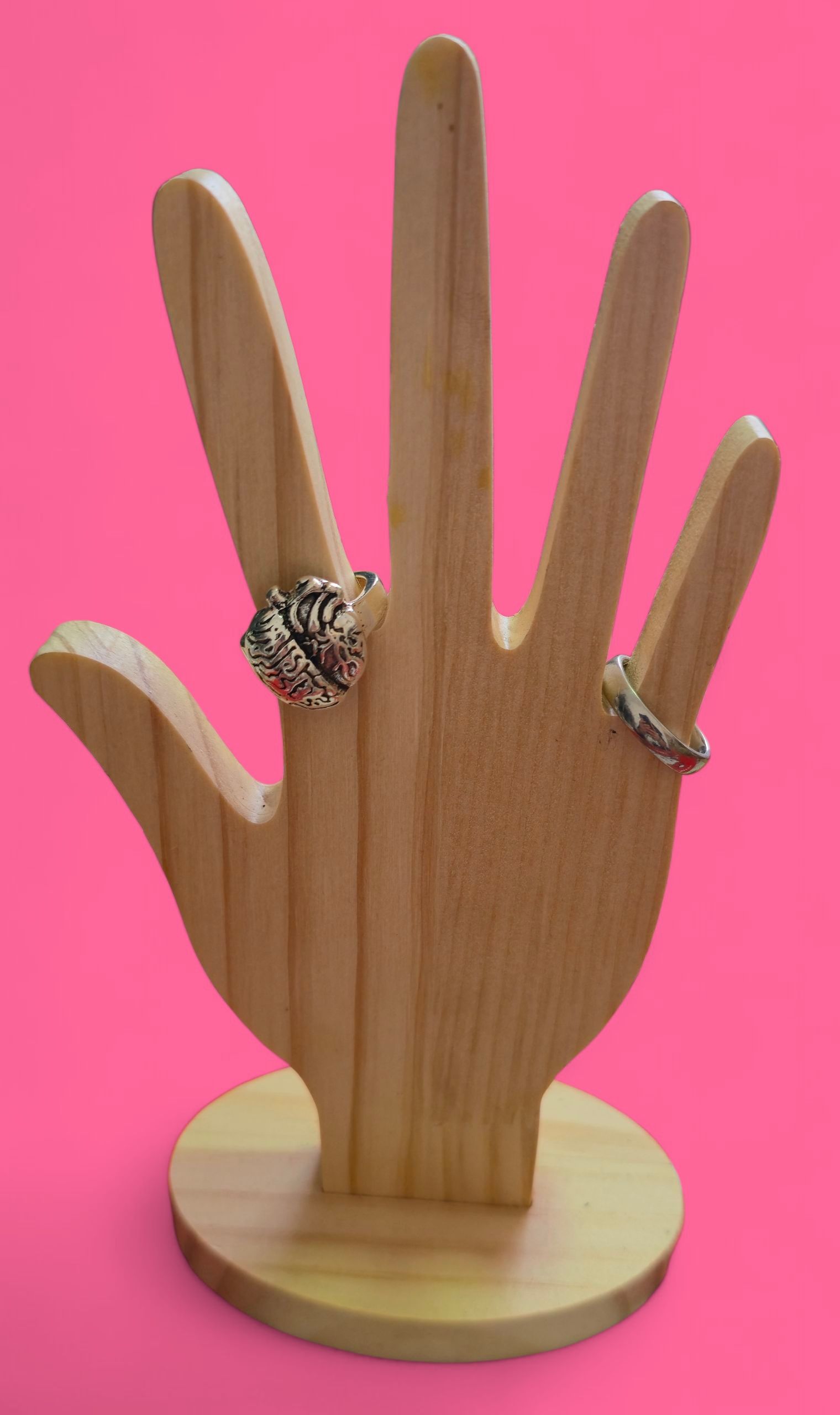 Anillo plateado en forma de corazón sobre una mano de madera con fondo rosa.