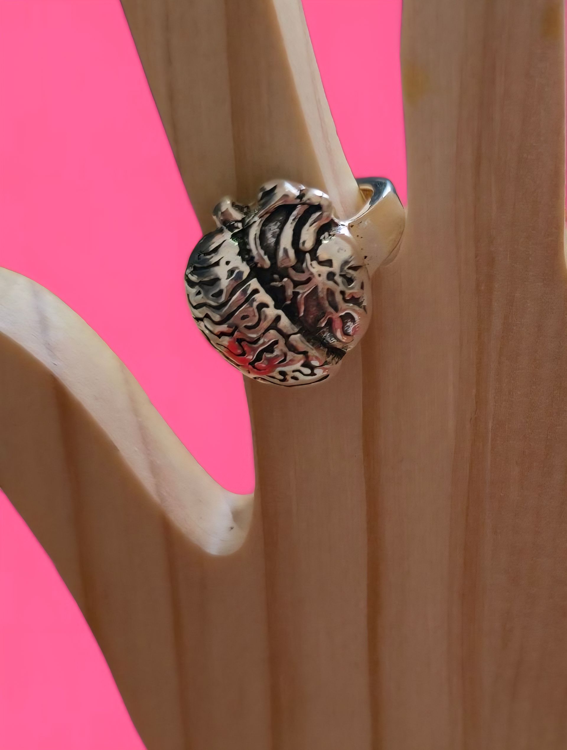 Anillo plateado en forma de corazón sobre una mano de madera con fondo rosa.