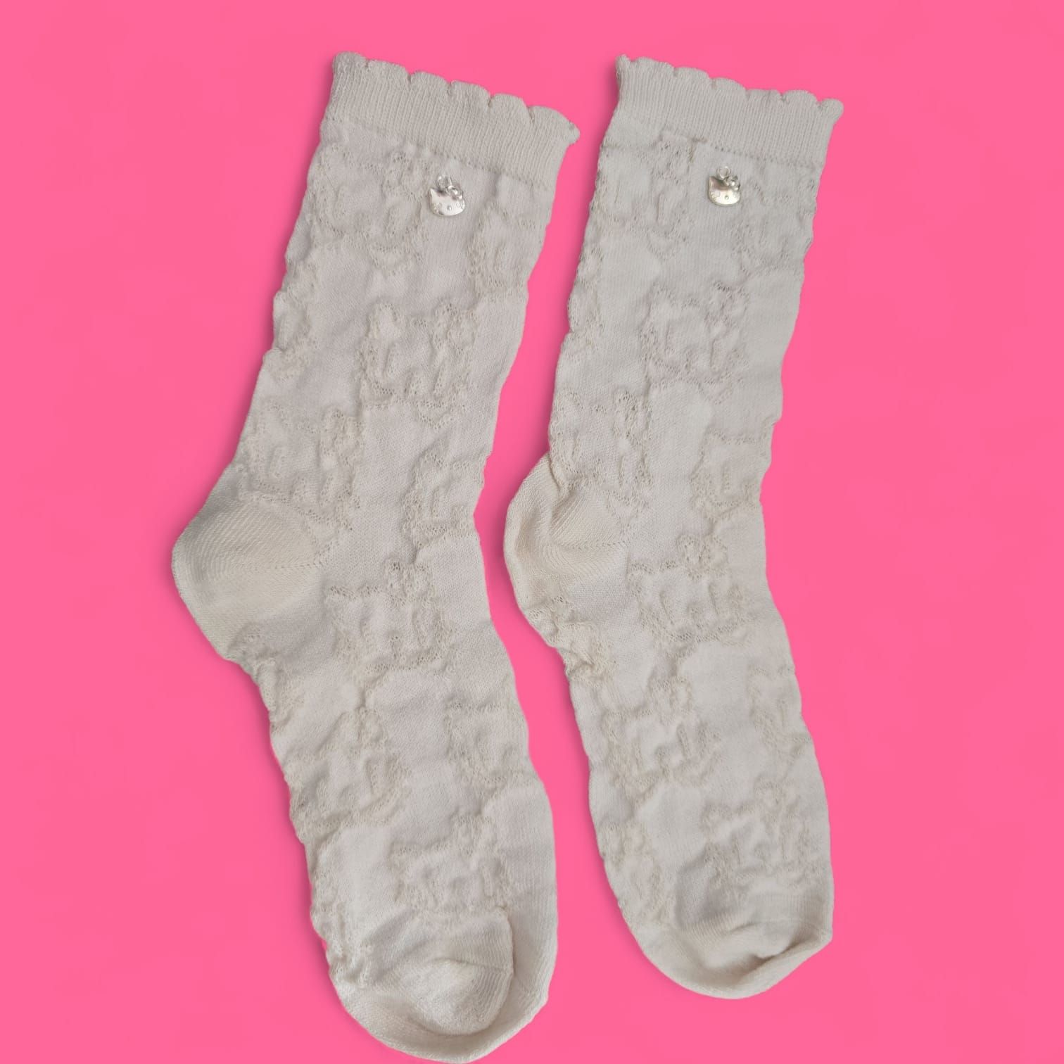 Dos calcetines de encaje color crudo con borde festoneado y un pequeño detalle de cuentas plateadas, sobre un fondo rosa.