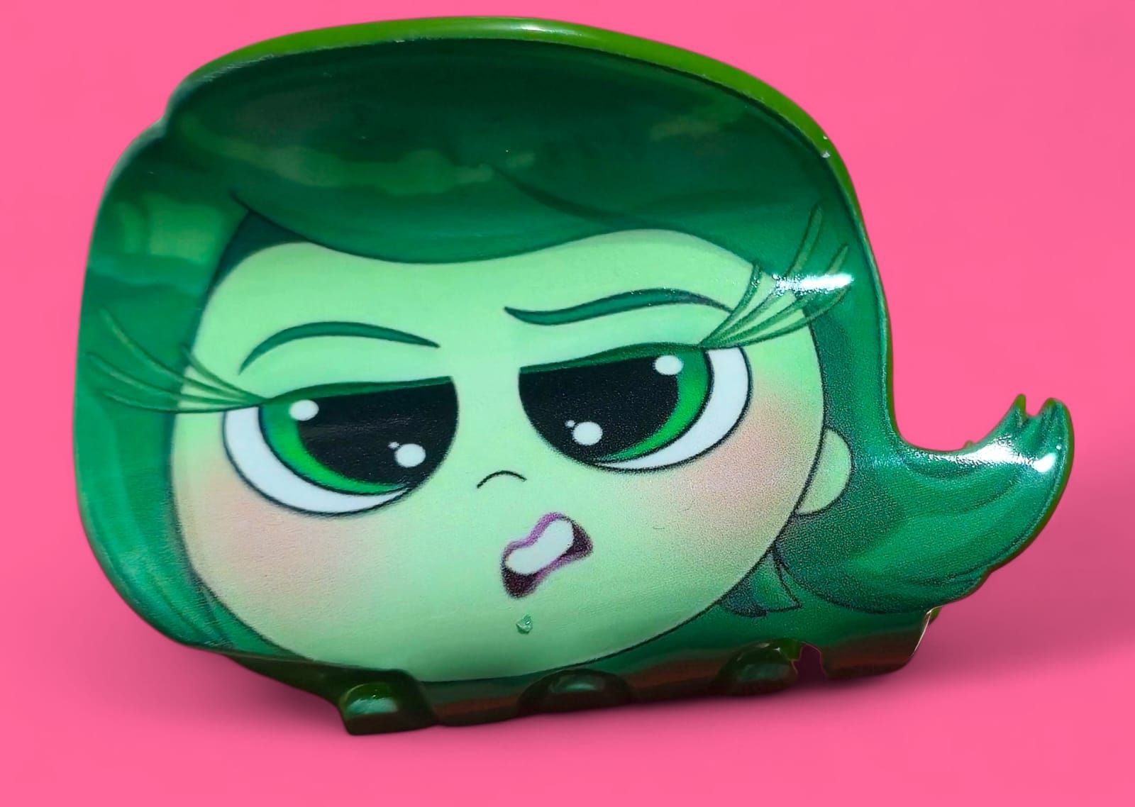 Primer plano de Asco de la película Inside Out, en color verde, con expresión de asco, sobre un fondo rosa.
