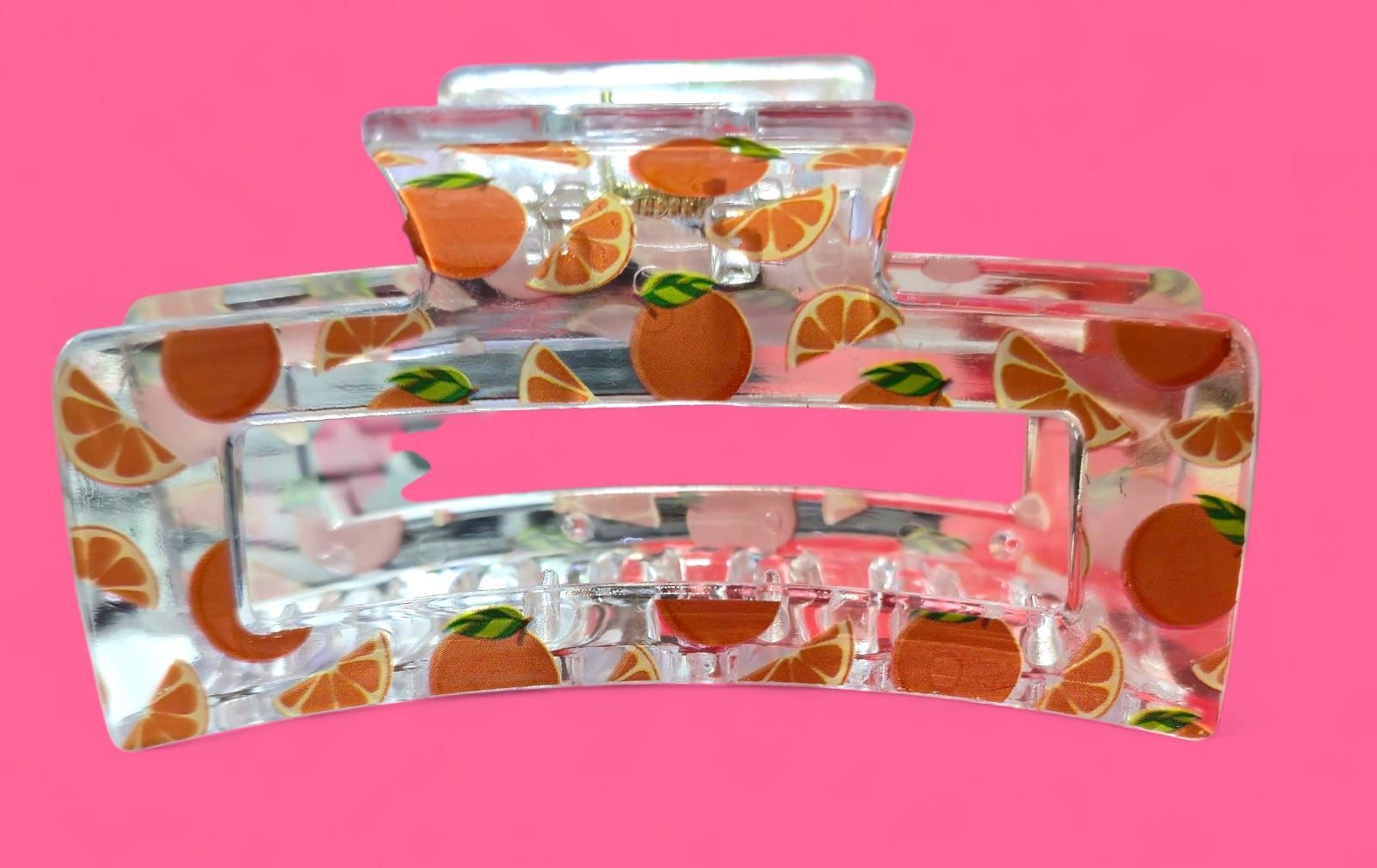 Pinza para el pelo de plástico transparente con rodajas de naranja y naranjas enteras sobre fondo rosa.