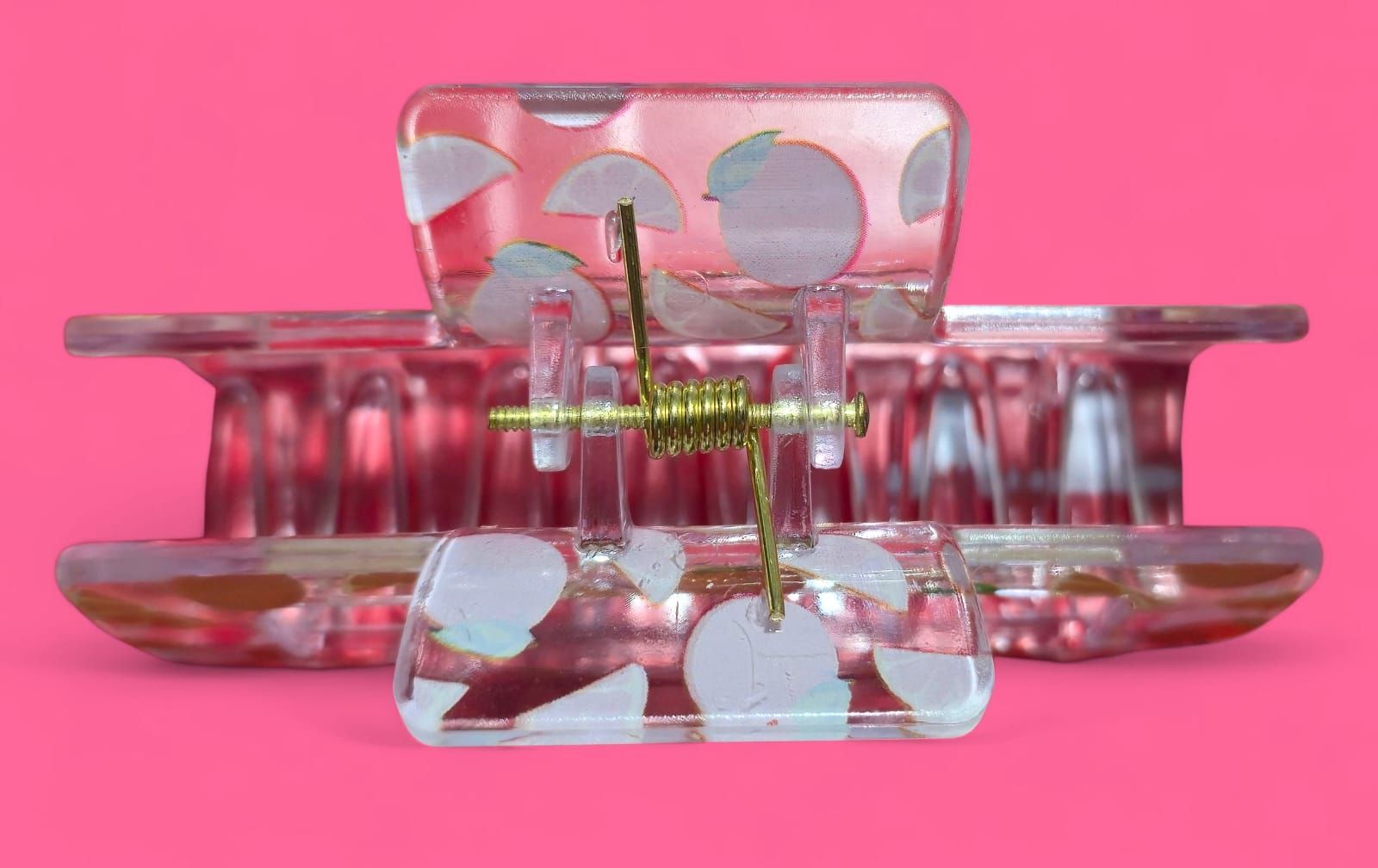 Pinza para el pelo de plástico transparente con rodajas de naranja y naranjas enteras sobre fondo rosa.