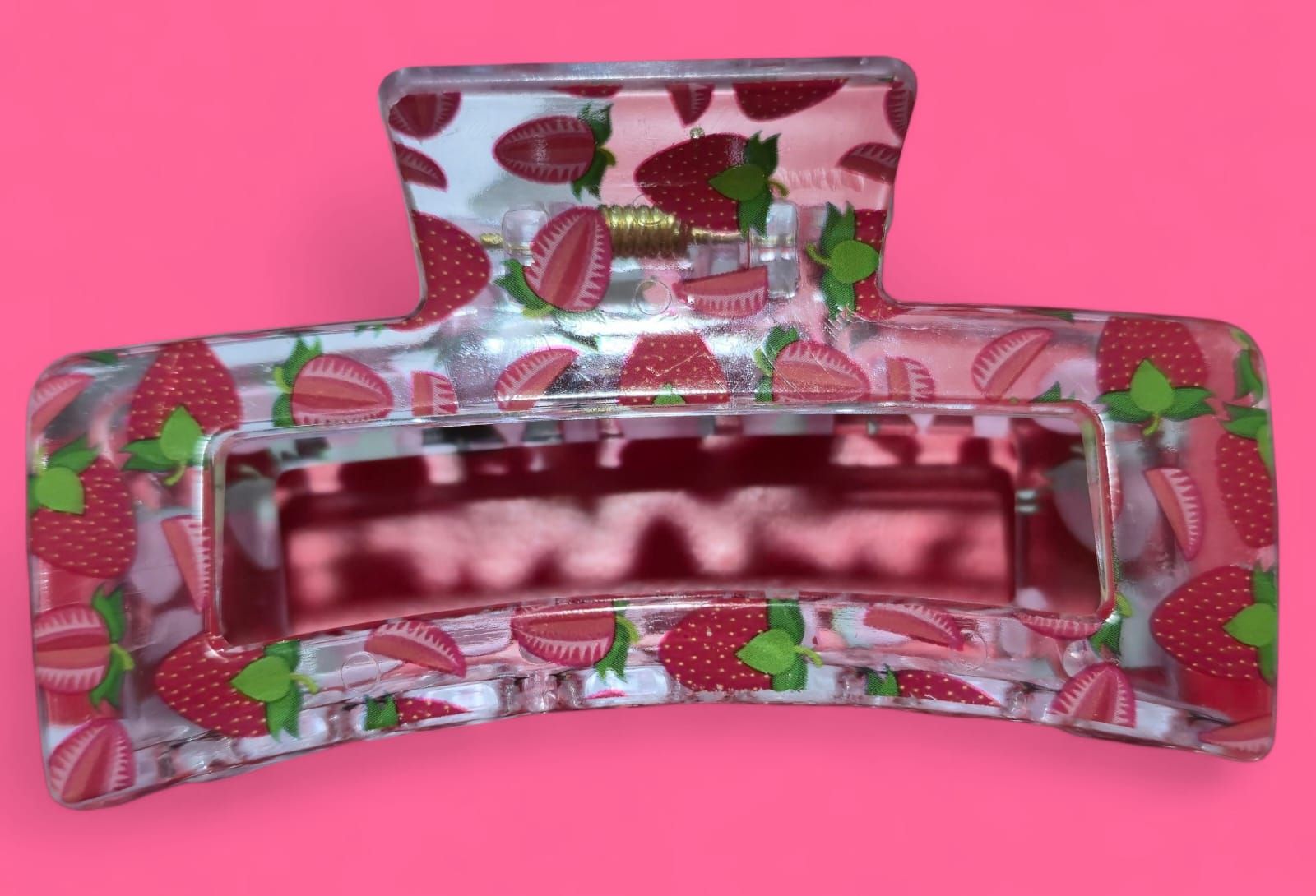 Pinza para el cabello con estampado de fresas, transparente con fresas rojas y verdes, sobre fondo rosa.