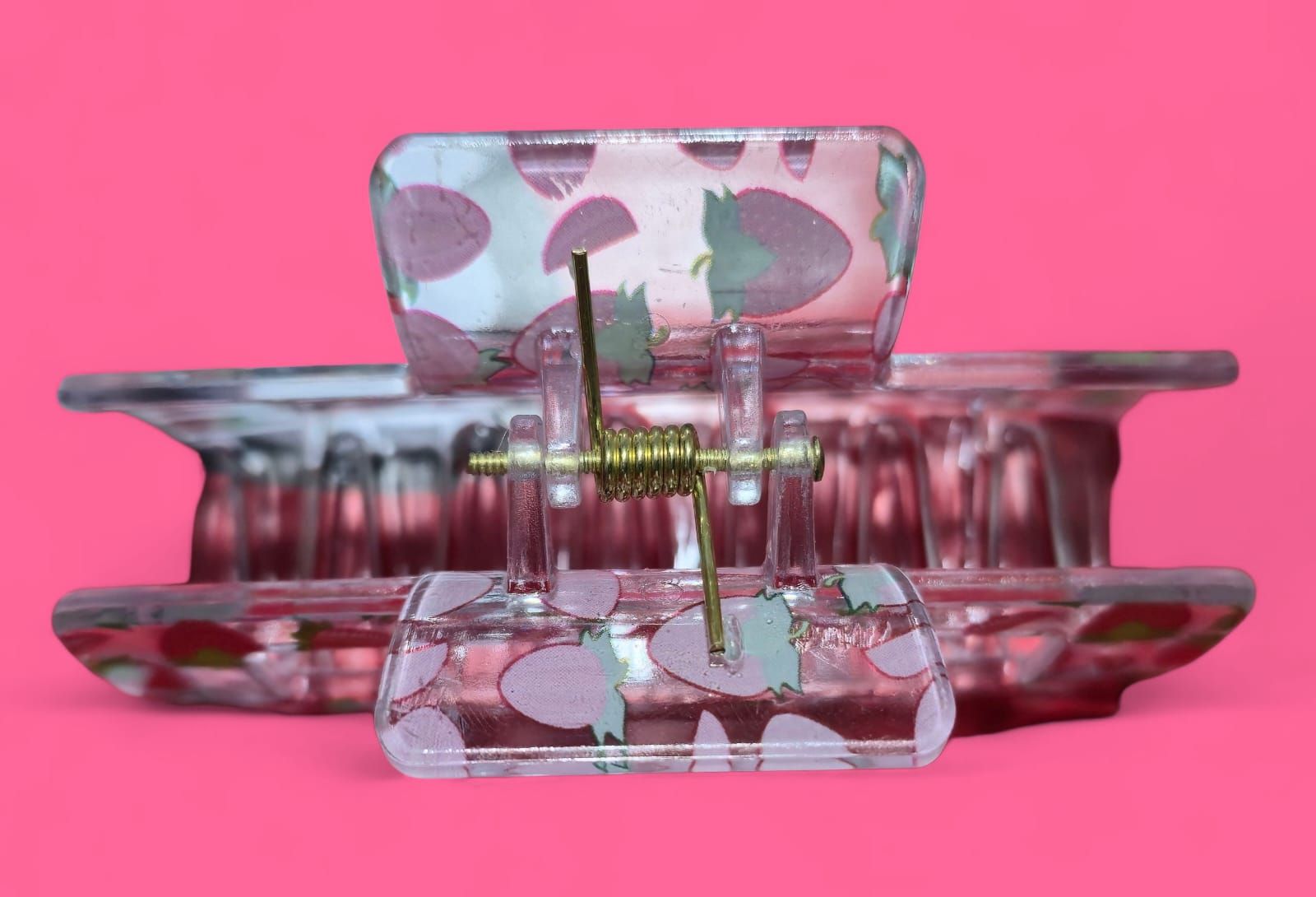 Pinza para el cabello con estampado de fresas, transparente con fresas rojas y verdes, sobre fondo rosa.