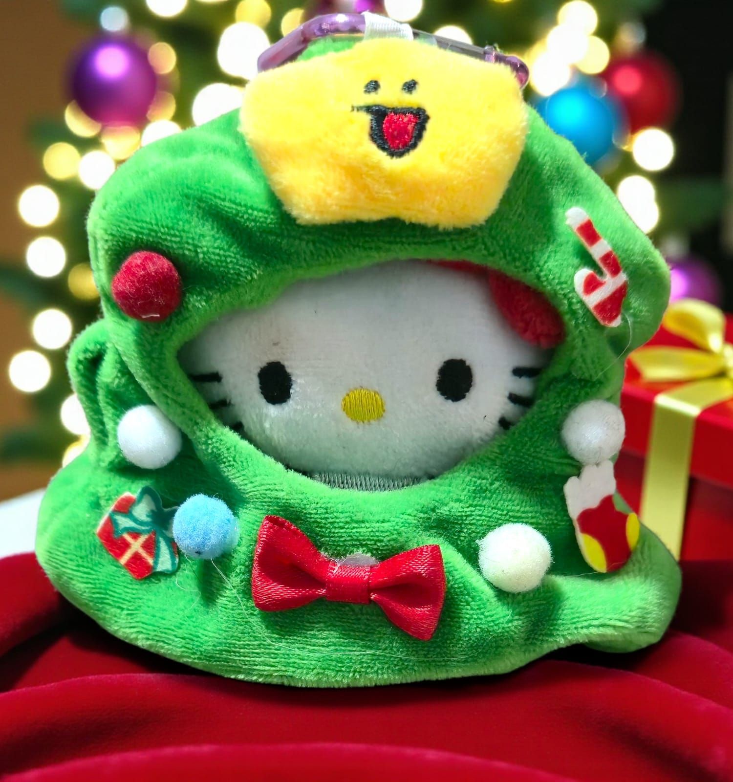 Peluche de Hello Kitty dentro de un disfraz verde de árbol de Navidad, con lazo rojo y adornos coloridos.