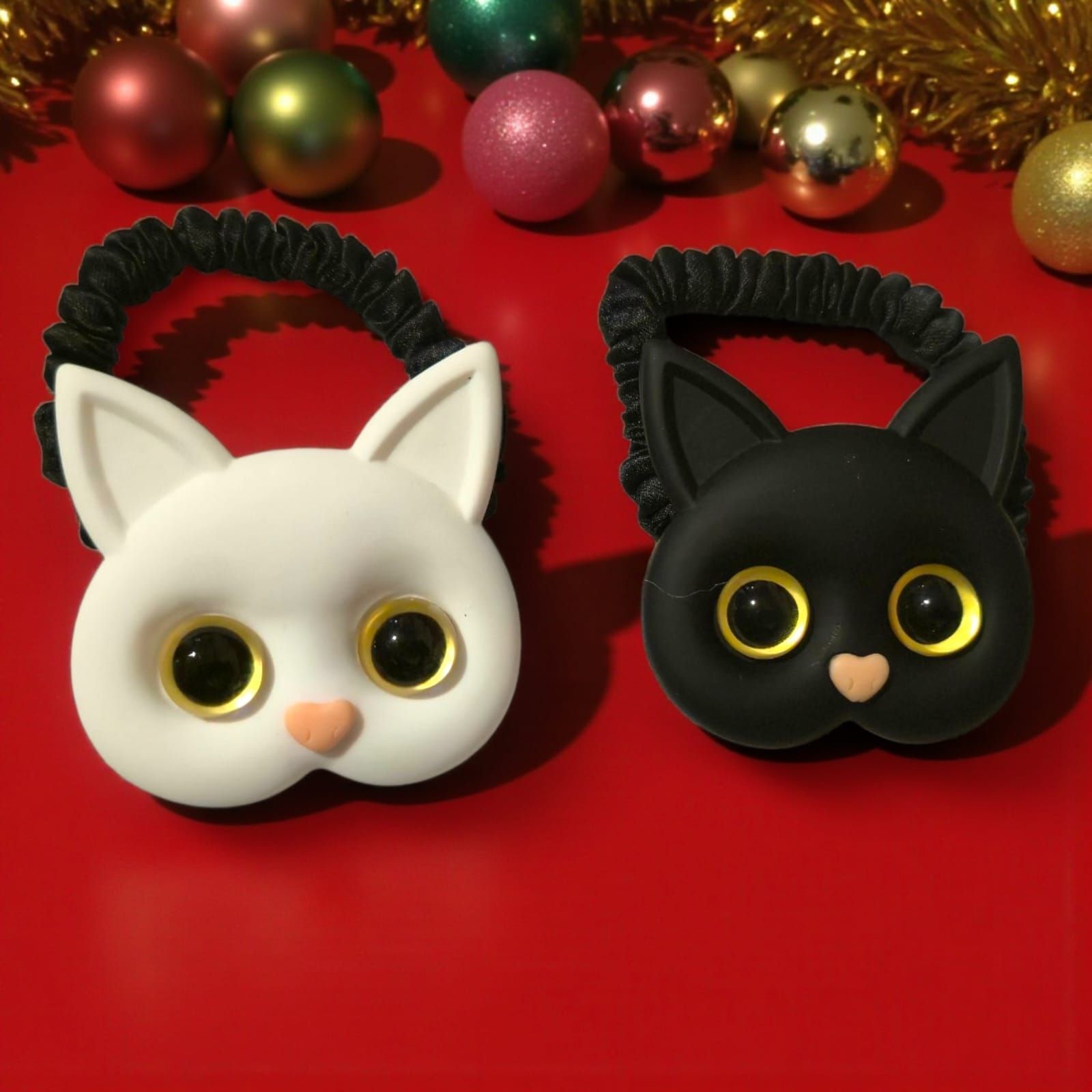 Dos gomas para el pelo con forma de gato: una blanca y otra negra, cada una con ojos saltones y nariz rosa.