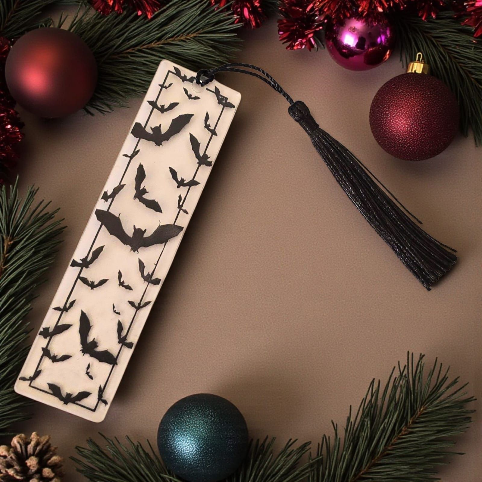 Marcapáginas con siluetas de murciélagos negros y una borla negra, rodeado de adornos navideños y vegetación.