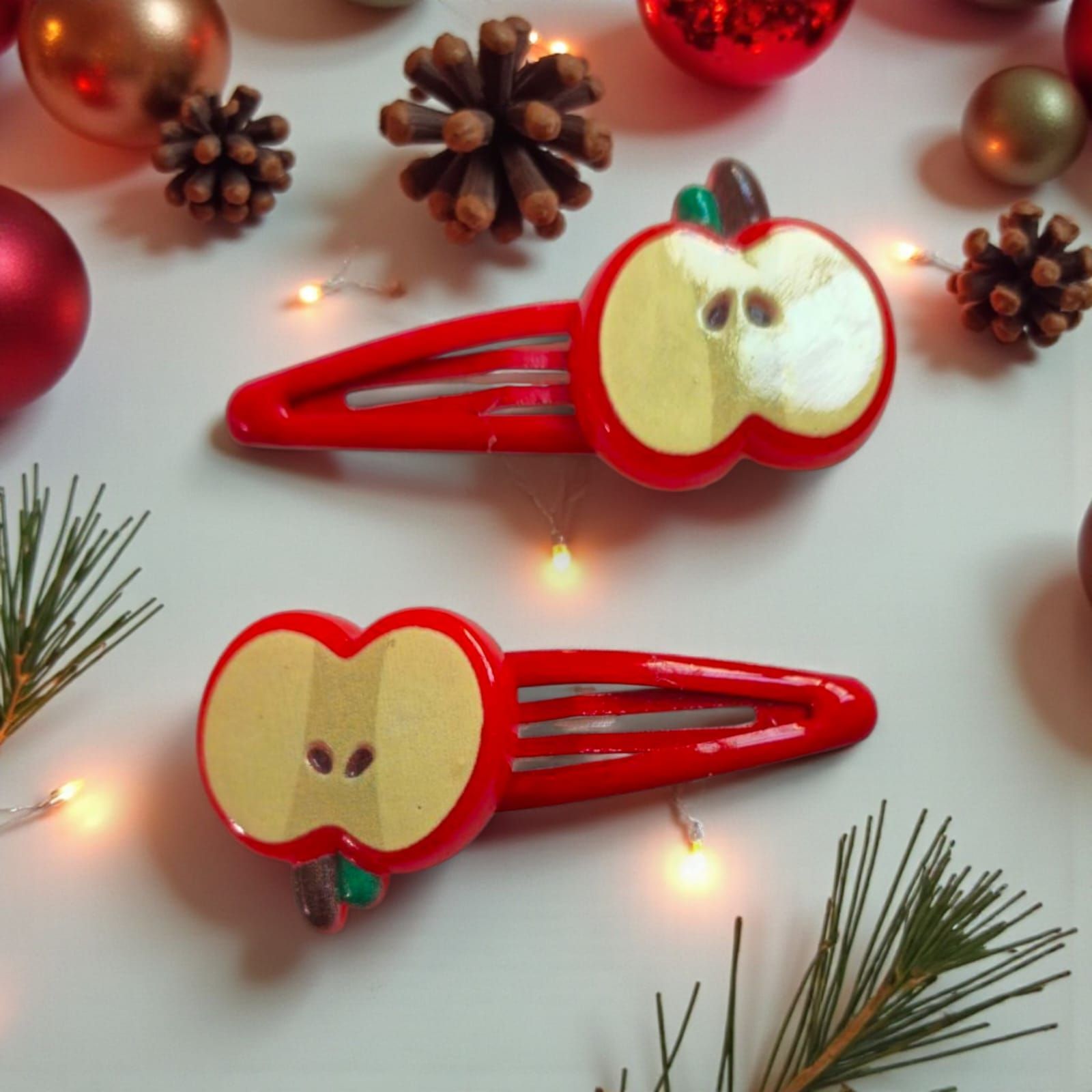 Dos pinzas para el cabello rojas con forma de manzana y centro beige, rodeadas de adornos navideños y agujas de pino.