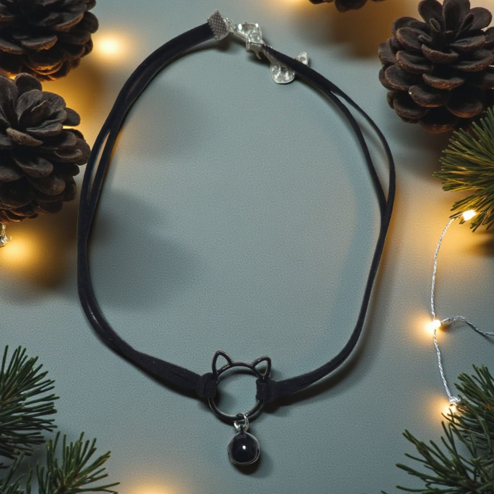 Gargantilla negra con colgante de cabeza de gato y una piedra negra sobre una superficie gris con piñas y luces.