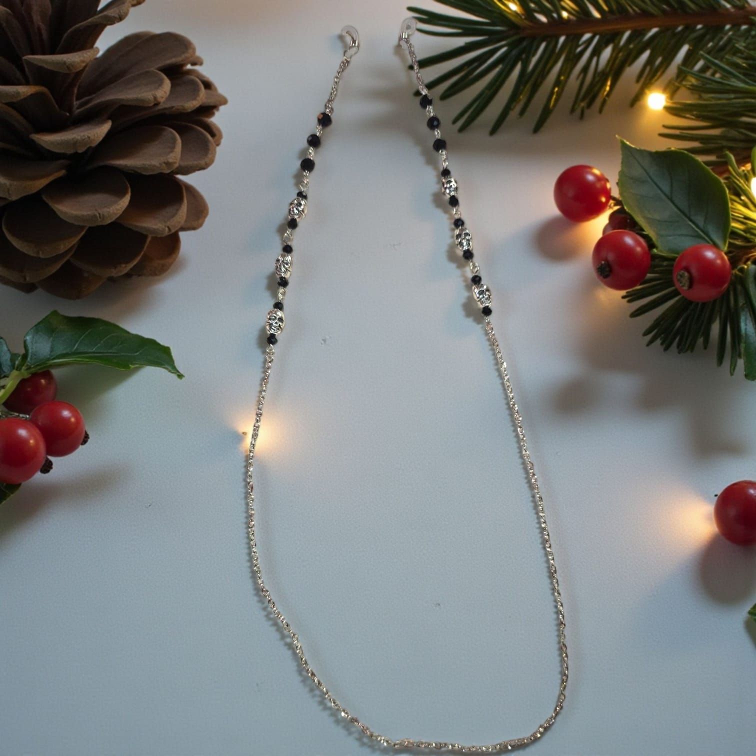 Cadena para gafas con cuentas negras y cadena plateada, junto a piñas, bayas y vegetación.