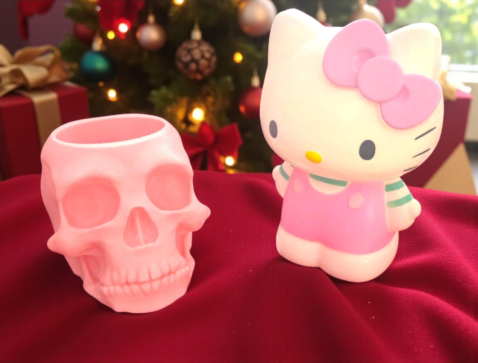 Maceta con forma de calavera rosa y figura de Hello Kitty sobre tela roja, árbol de Navidad al fondo.