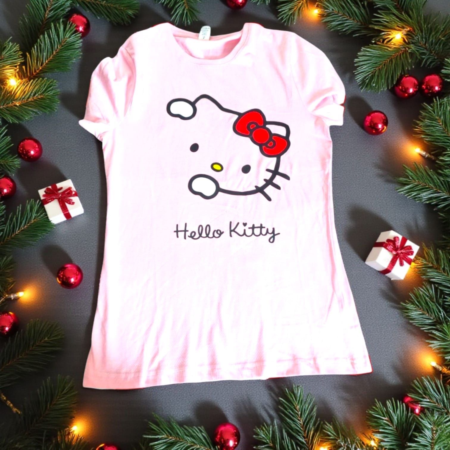 Camiseta rosa de Hello Kitty con un lazo rojo, rodeada de adornos navideños y vegetación.