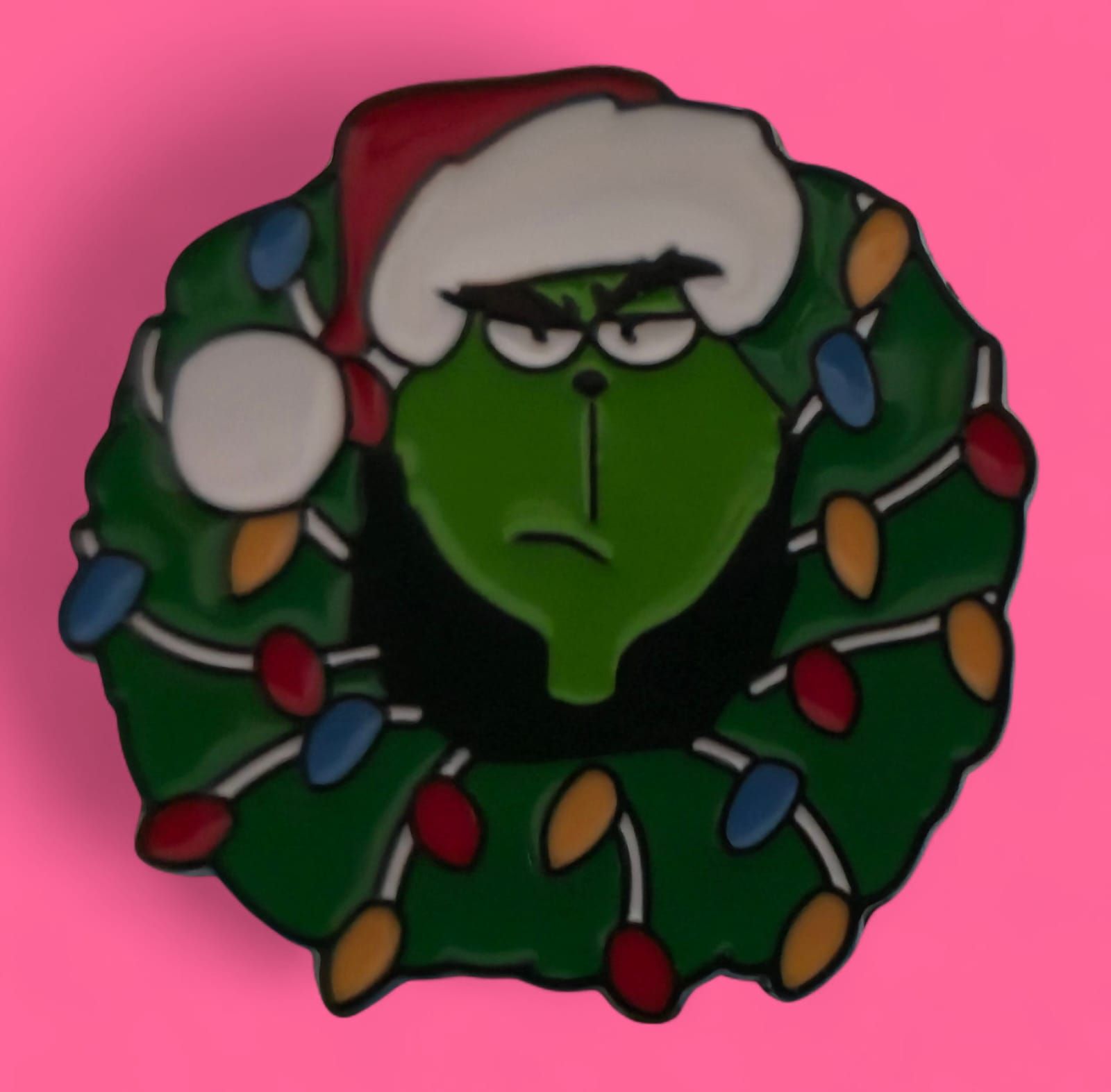 El Grinch con gorro de Papá Noel en una corona verde con luces navideñas, sobre un fondo rosa.
