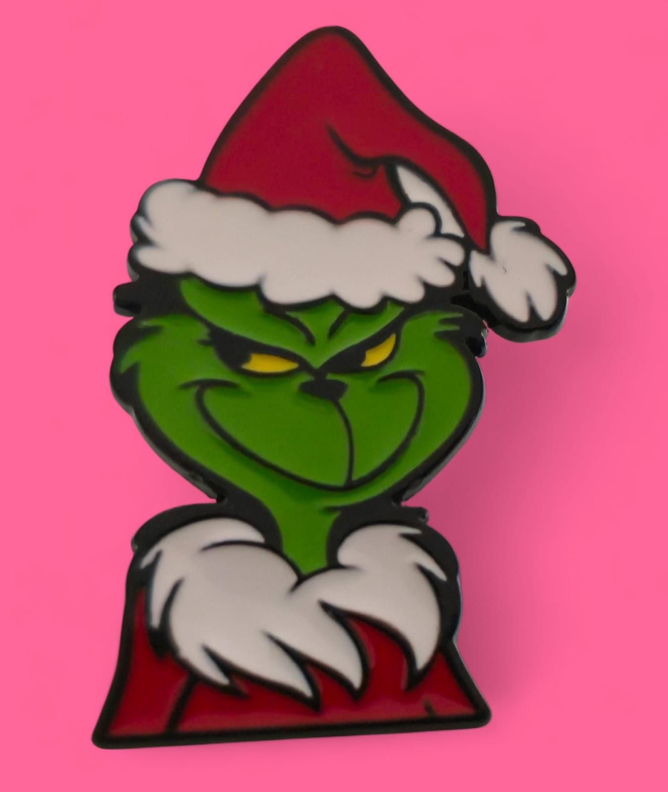 El Grinch con gorro de Papá Noel, sonriendo con ojos amarillos, sobre un fondo rosa.