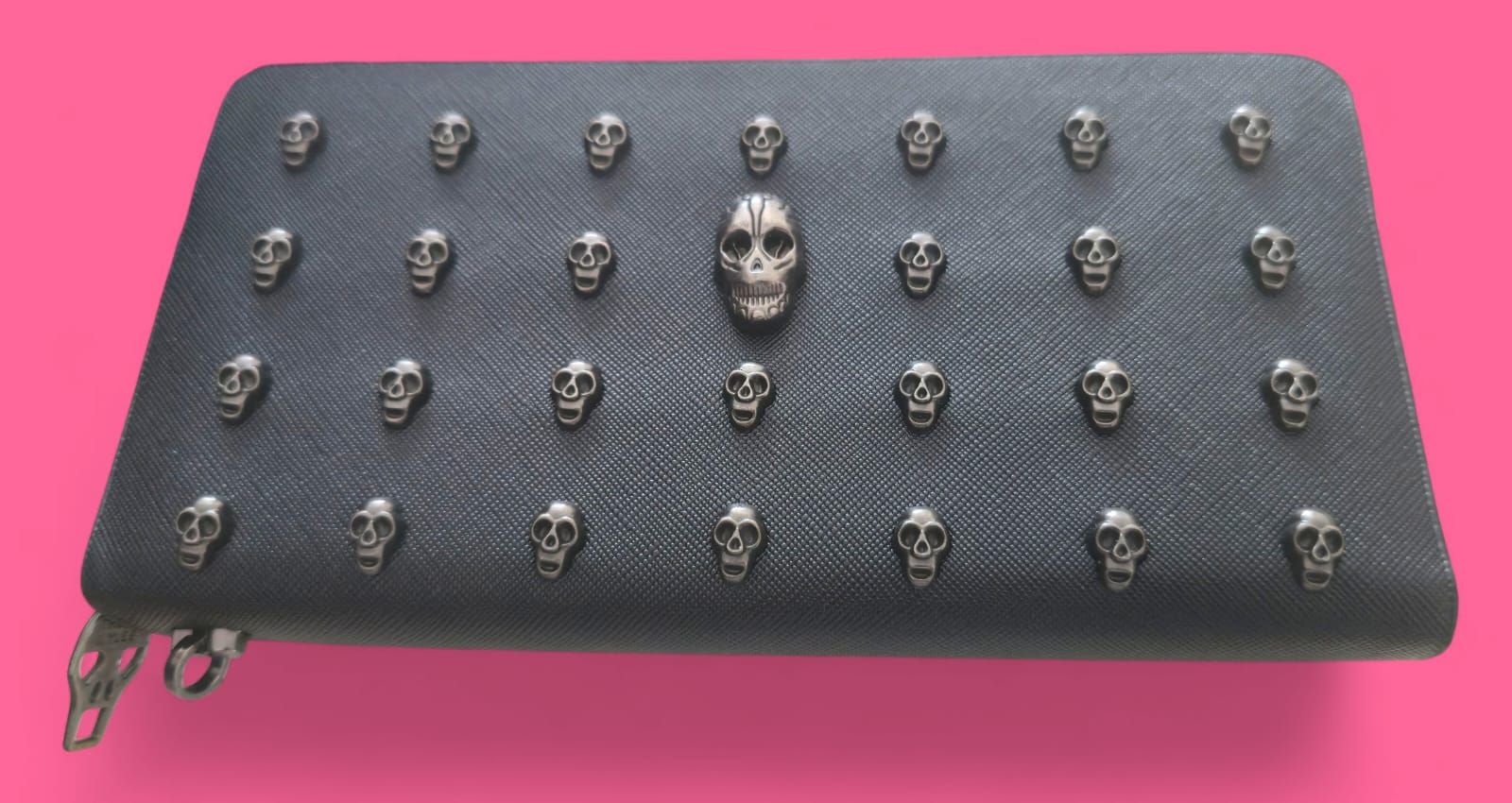 Cartera negra con tachuelas de calavera, calavera central más grande, cremallera plateada, fondo rosa.