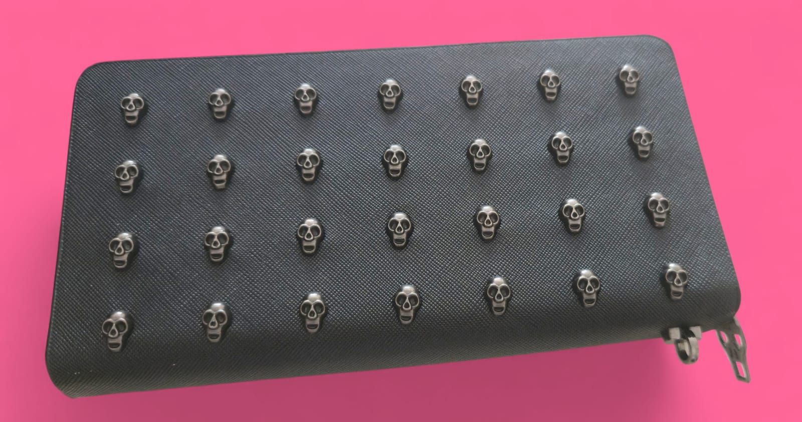 Cartera negra con tachuelas de calavera, calavera central más grande, cremallera plateada, fondo rosa.