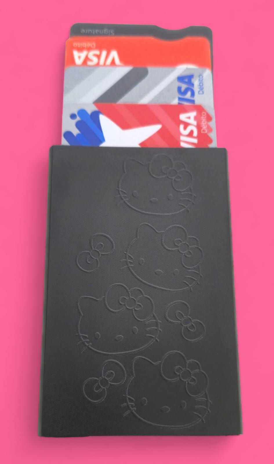 Caja rectangular negra con caras y lazos de Hello Kitty en relieve. Fondo rosa.