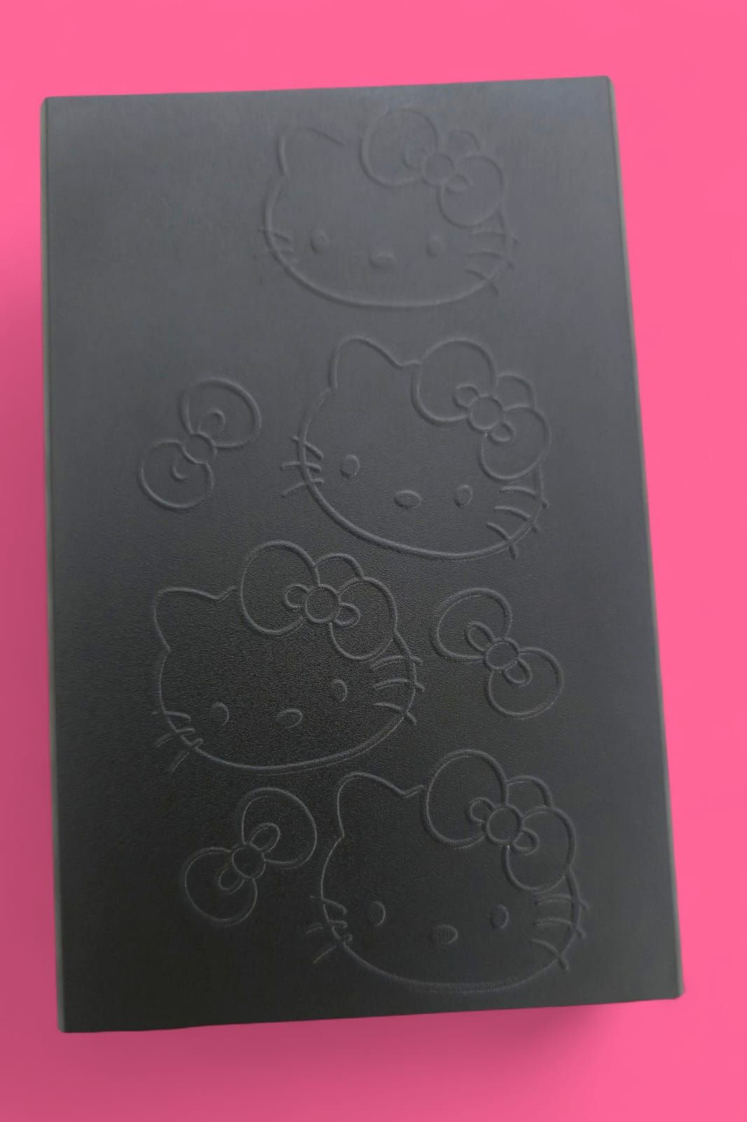 Caja rectangular negra con caras y lazos de Hello Kitty en relieve. Fondo rosa.