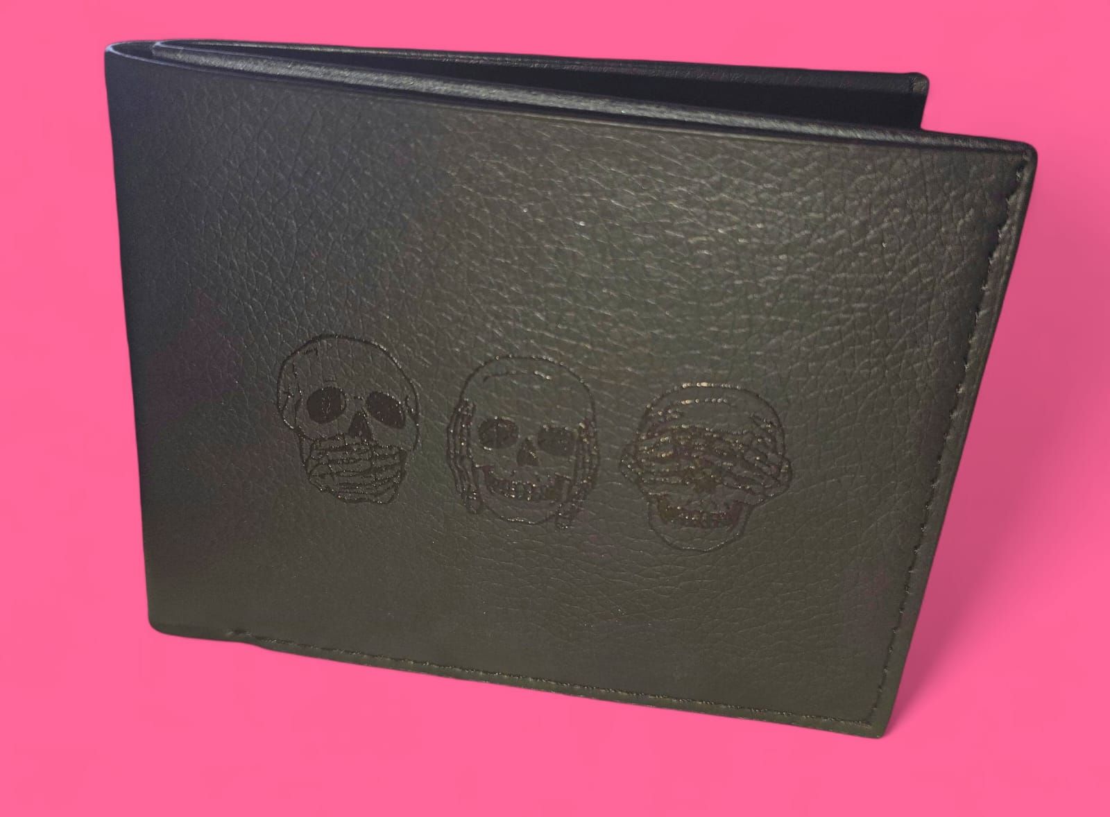 Cartera de piel negra con tres diseños de calaveras sobre fondo rosa.