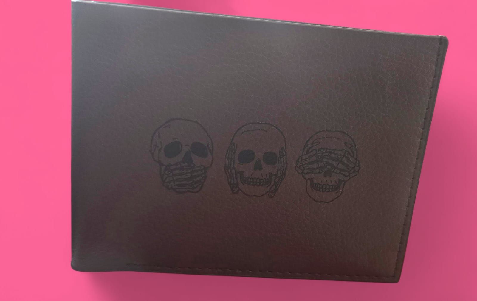 Cartera de piel marrón con tres diseños de calaveras sobre fondo rosa.