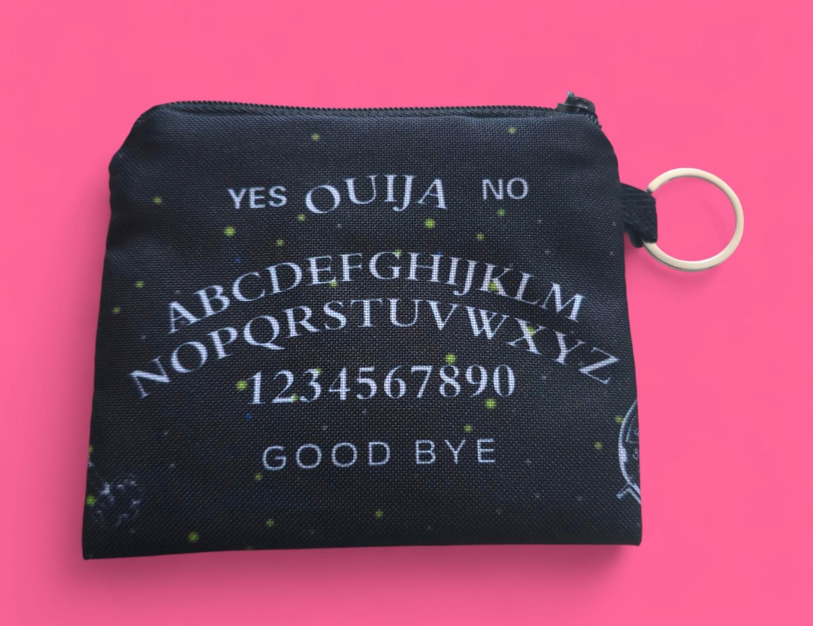 Monedero negro con estampado de tablero Ouija, texto blanco y cremallera dorada sobre fondo rosa.