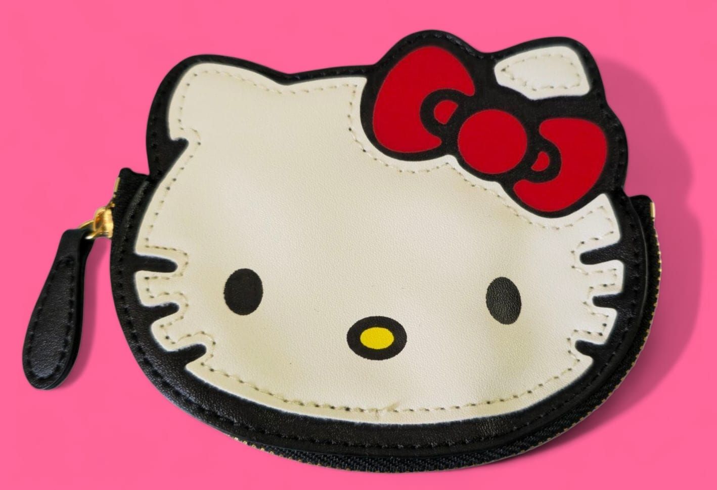 Monedero Hello Kitty, cara color crema con contorno negro, lazo rojo, cremallera negra sobre fondo rosa.