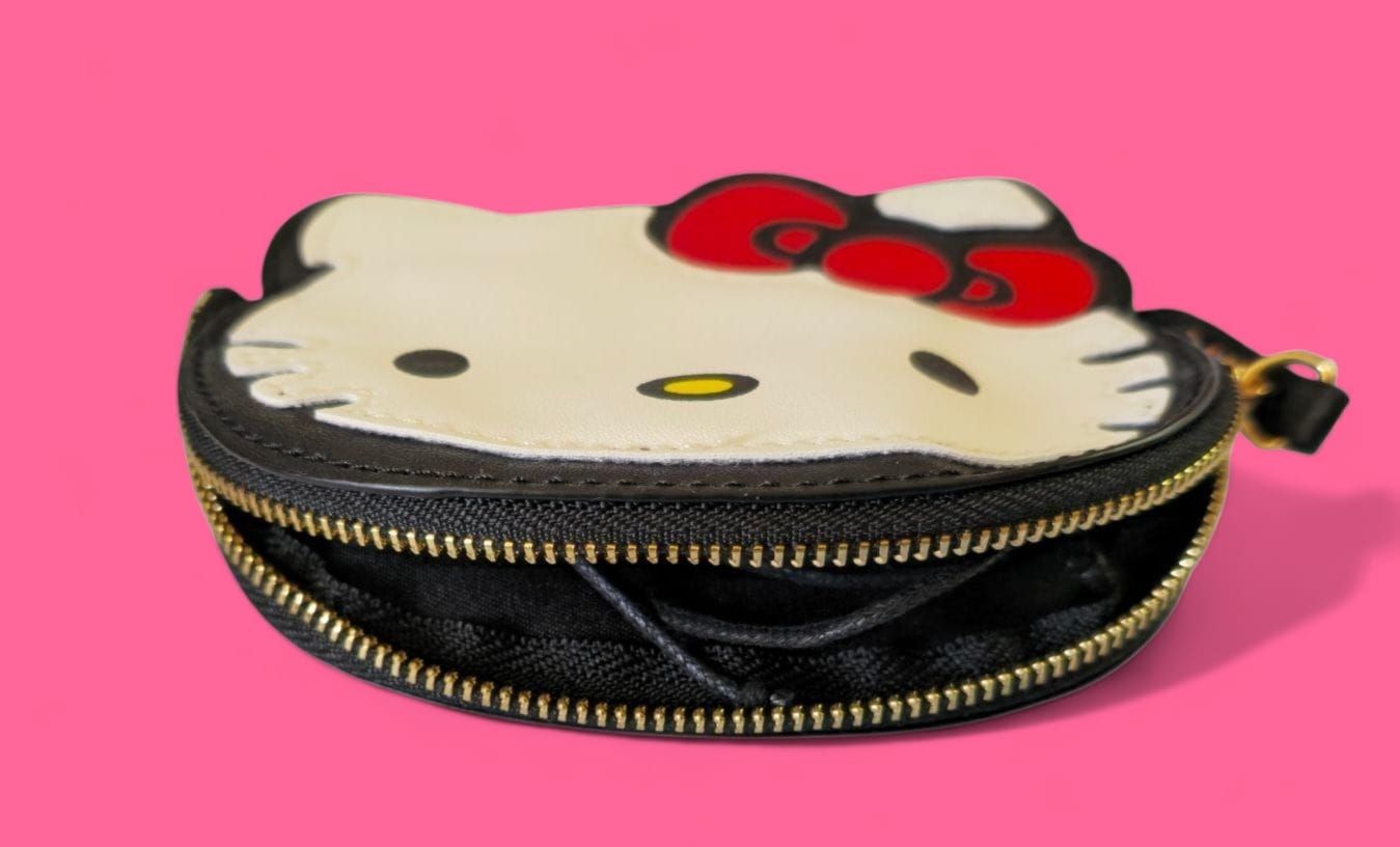 Monedero Hello Kitty, cara color crema con contorno negro, lazo rojo, cremallera negra sobre fondo rosa.