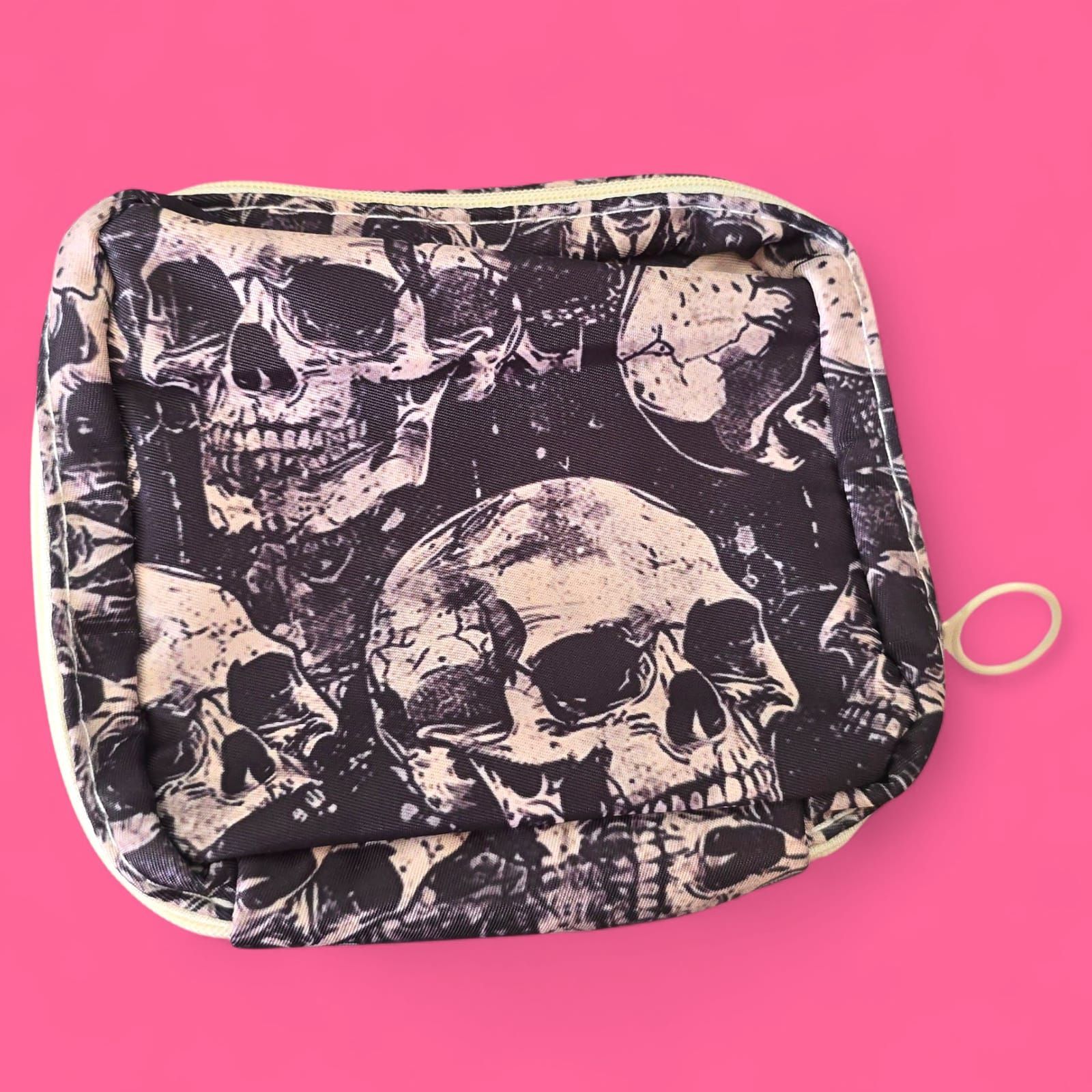 Neceser con estampado de calaveras, calaveras negras y crema, cremallera blanca, sobre fondo rosa.