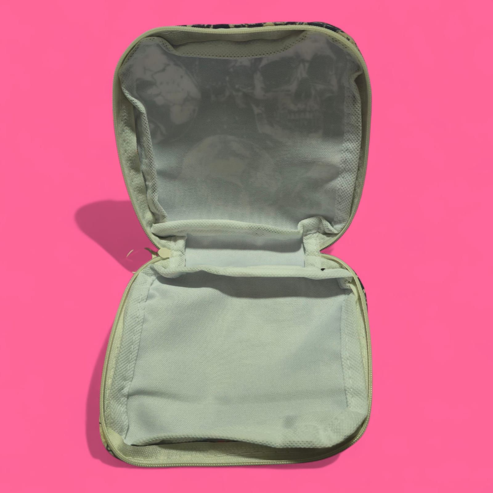 Neceser con estampado de calaveras, calaveras negras y crema, cremallera blanca, sobre fondo rosa.