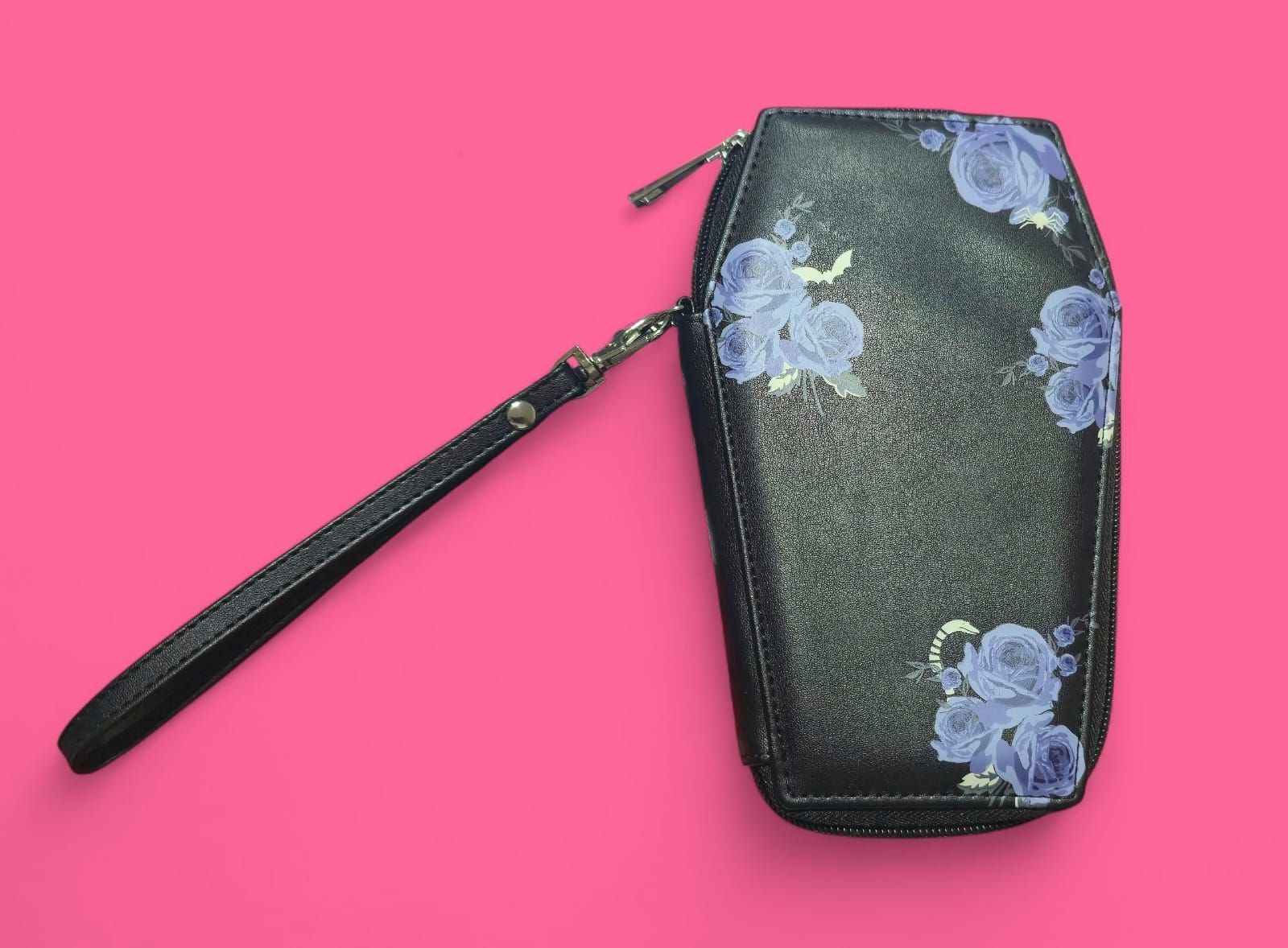Cartera negra con forma de ataúd, diseño floral azul y correa de muñeca sobre fondo rosa.