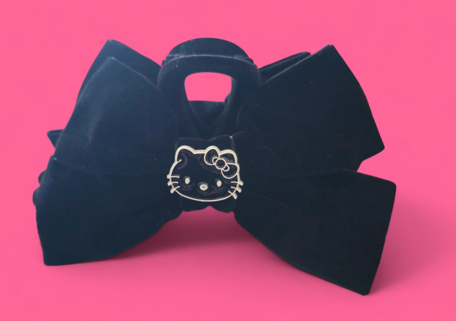 Pinza para el pelo con lazo de terciopelo negro y adorno de Hello Kitty sobre fondo rosa.