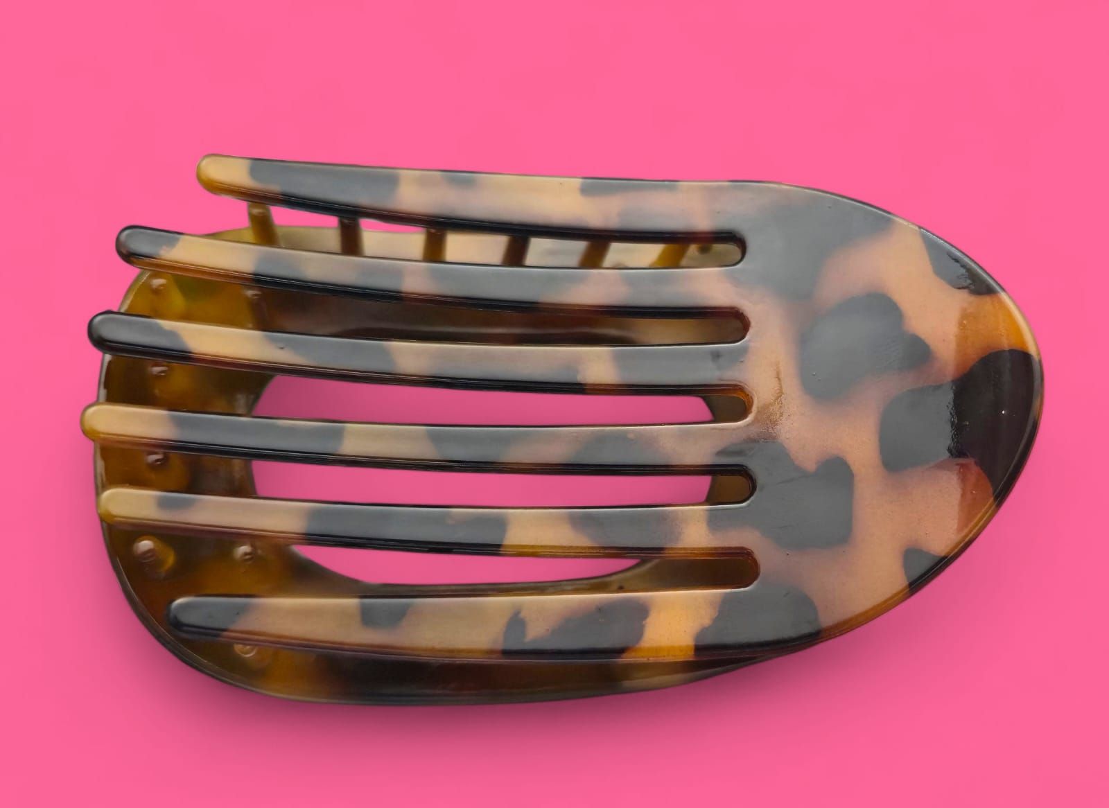 Pinza para el cabello con estampado de carey sobre fondo rosa, de forma ovalada con listones horizontales.