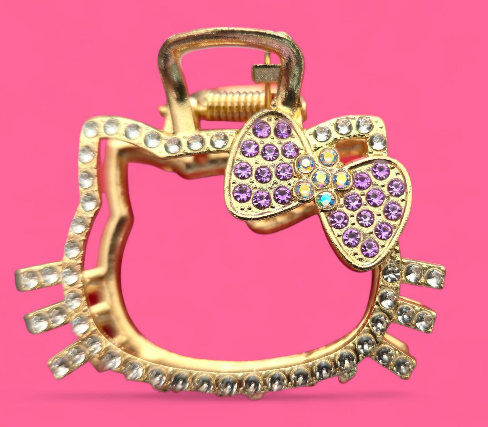 Pinza para el pelo Hello Kitty dorada con brillantes morados y transparentes sobre fondo rosa.