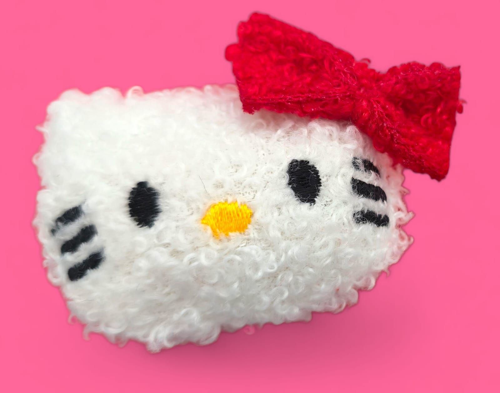 Peluche de Hello Kitty con forma de sushi, con lazo rojo, ojos negros, bigotes y pico amarillo, sobre fondo rosa.