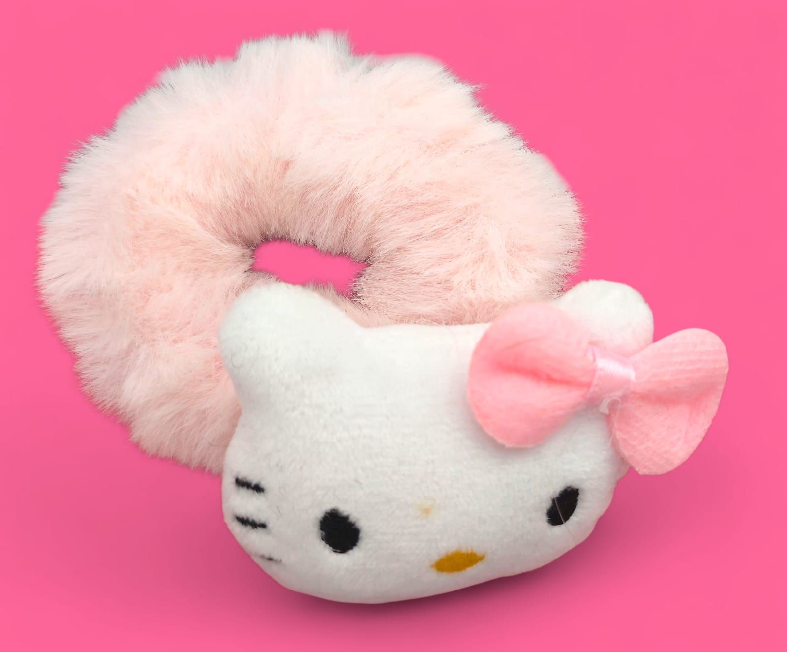 Coletero de Hello Kitty: rosa esponjoso, cara de peluche blanca, lazo rosa, sobre fondo rosa.