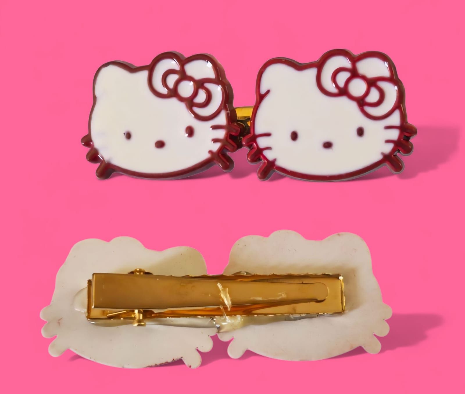 Dos horquillas de Hello Kitty sobre fondo rosa. Caritas blancas con lazos y contornos rojos. Pinza dorada.