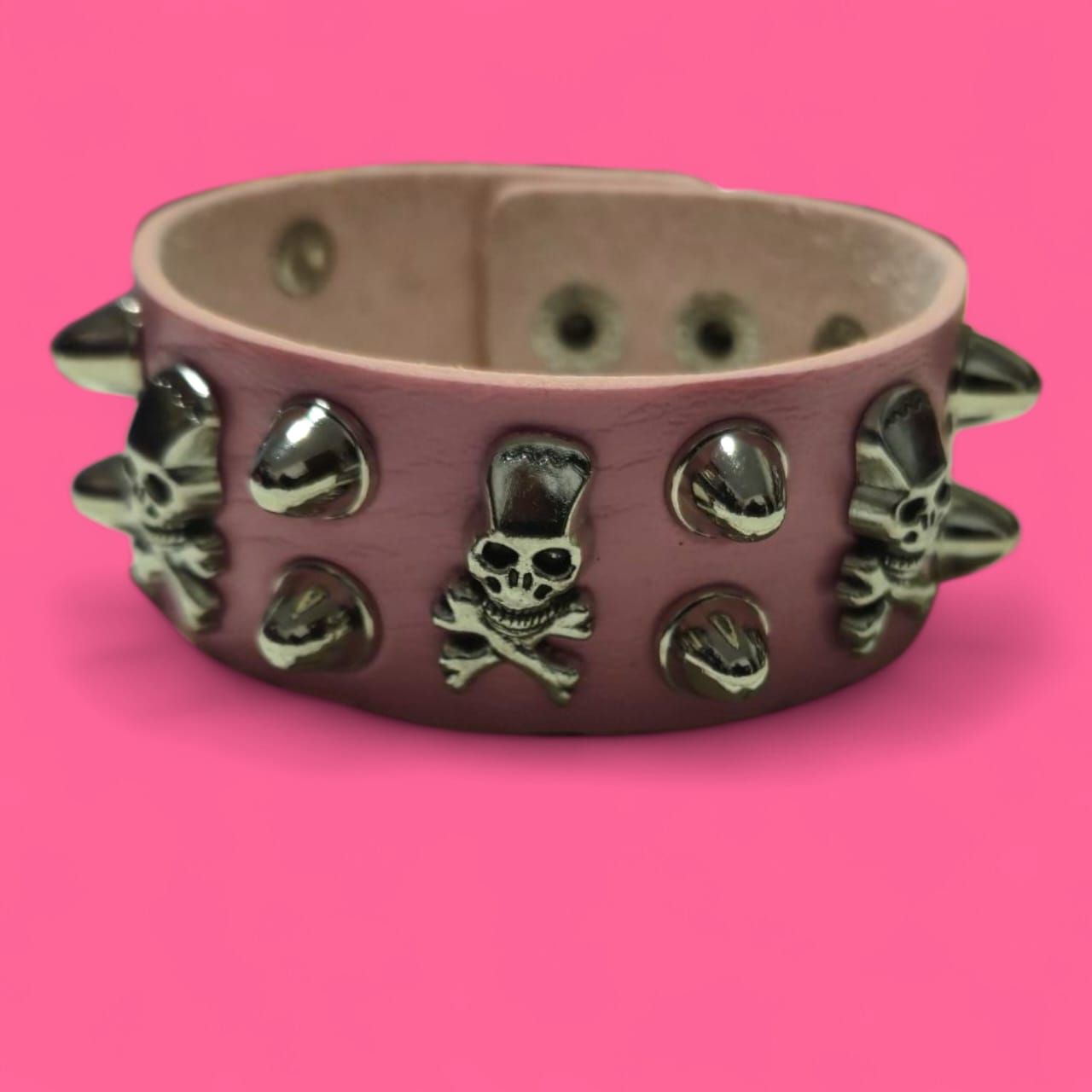 Pulsera rosa con tachuelas y remaches plateados en forma de calavera sobre fondo rosa.
