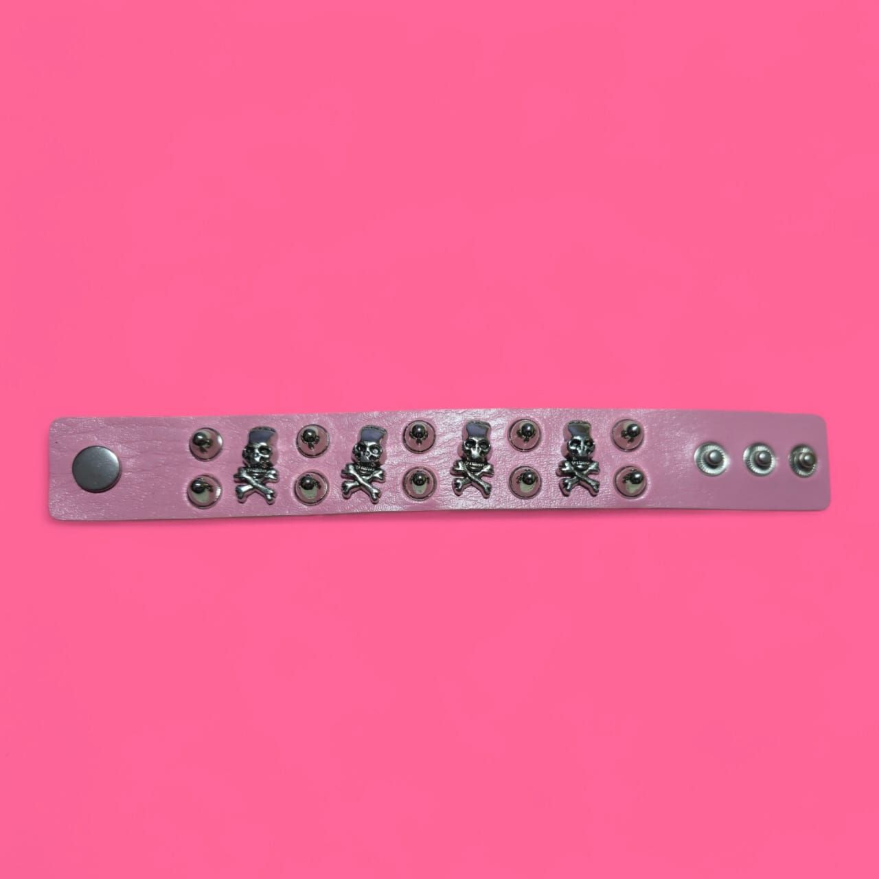 Pulsera rosa con tachuelas y remaches plateados en forma de calavera sobre fondo rosa.