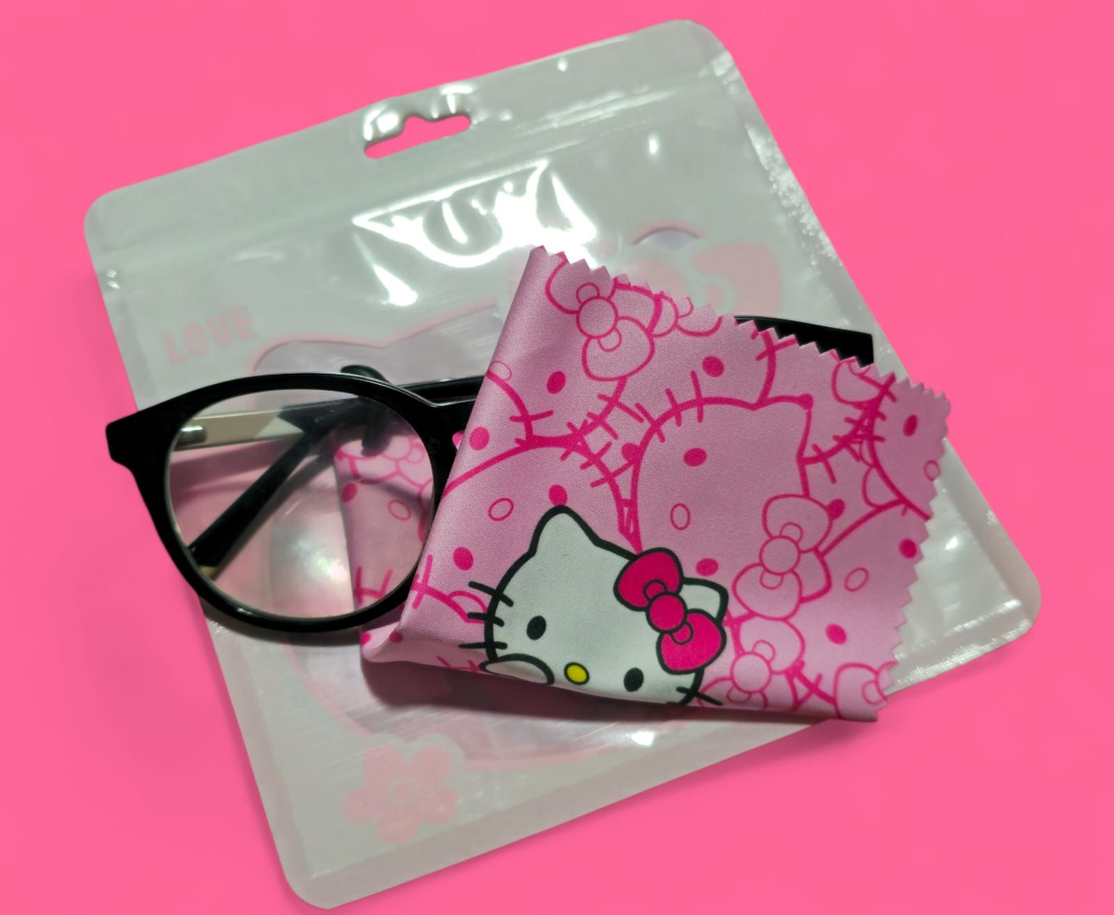 Gafas negras, paño de limpieza rosa de Hello Kitty y bolsa transparente sobre una superficie rosa.
