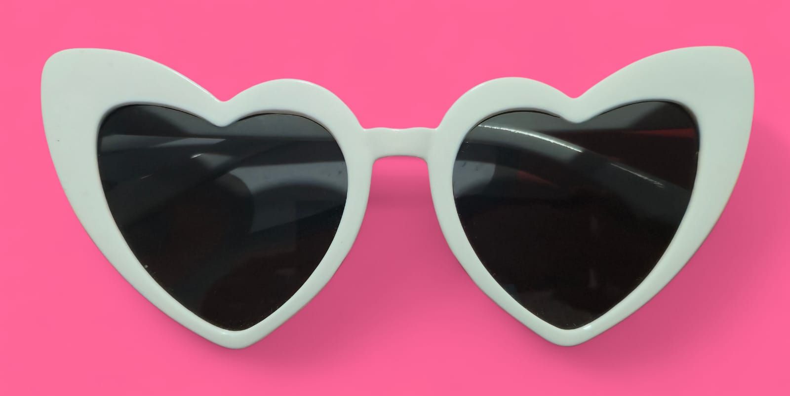 Gafas de sol blancas en forma de corazón con lentes oscuras sobre fondo rosa.
