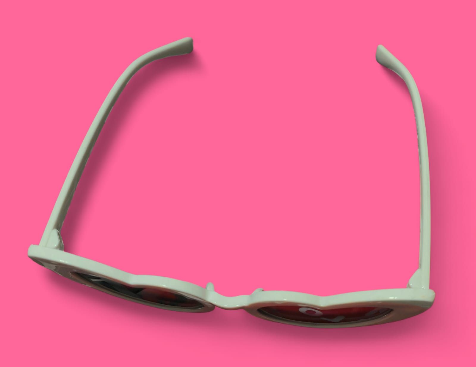 Gafas de sol blancas en forma de corazón con lentes oscuras sobre fondo rosa.