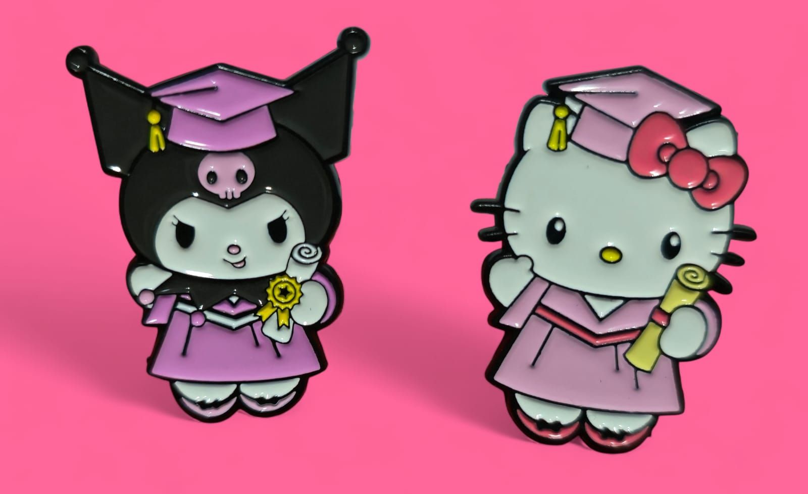 Pines de Kuromi y Hello Kitty con togas de graduación, sosteniendo diplomas, sobre fondo rosa.
