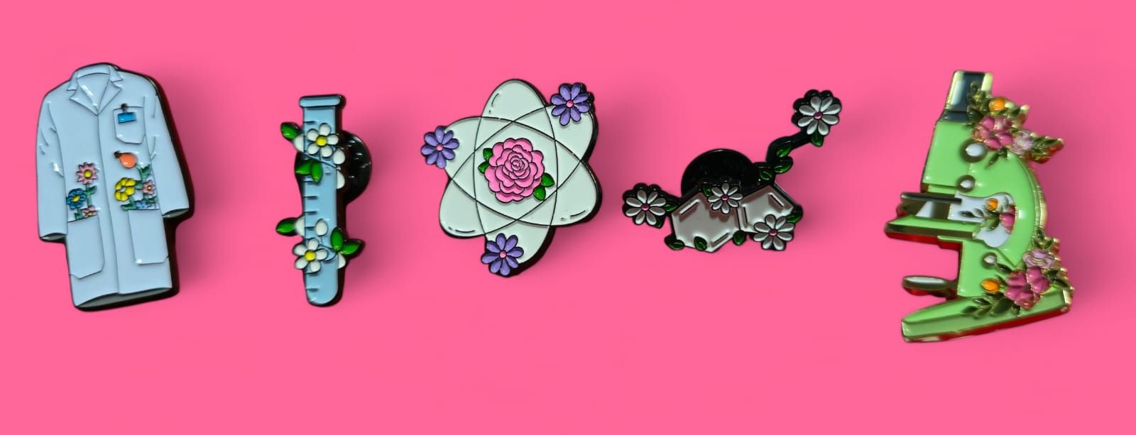 Cinco pins esmaltados sobre fondo rosa: bata de laboratorio, tubo de ensayo, átomo, gato y microscopio.