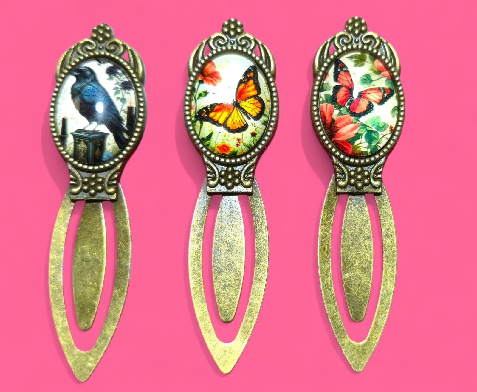 Tres marcadores de bronce con tapas ornamentadas: cuervo, mariposa monarca y mariposas rojas.
