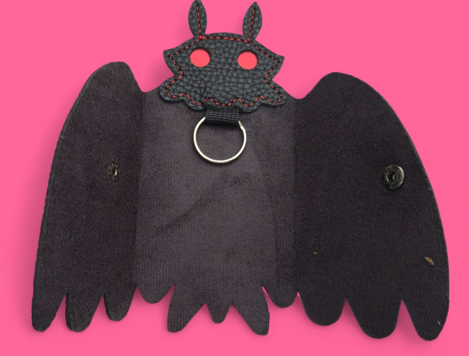 Estuche para llaves de piel de color negro con forma de Mothman, ojos rojos y cierre de botón, sobre fondo rosa.