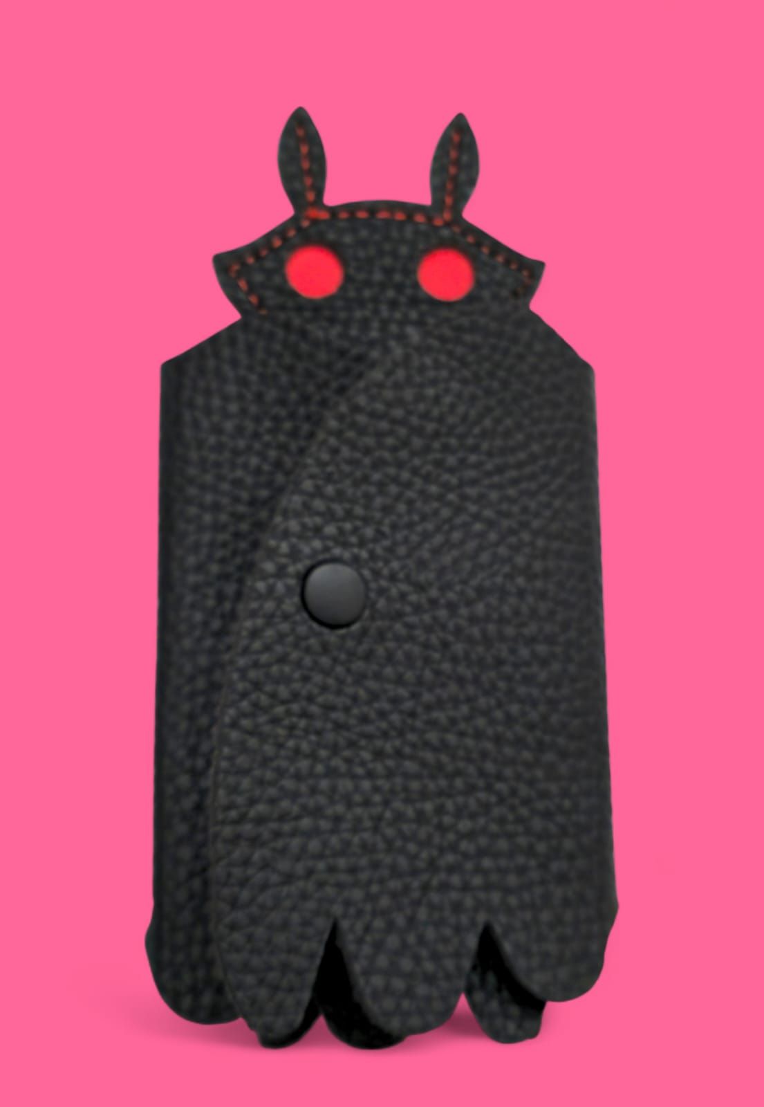 Estuche para llaves de piel de color negro con forma de Mothman, ojos rojos y cierre de botón, sobre fondo rosa.