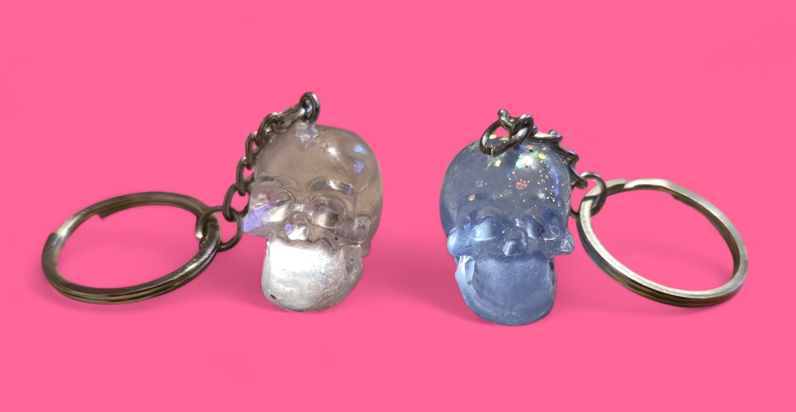 Dos llaveros con forma de calavera, uno plateado y otro azul, sobre fondo rosa.
