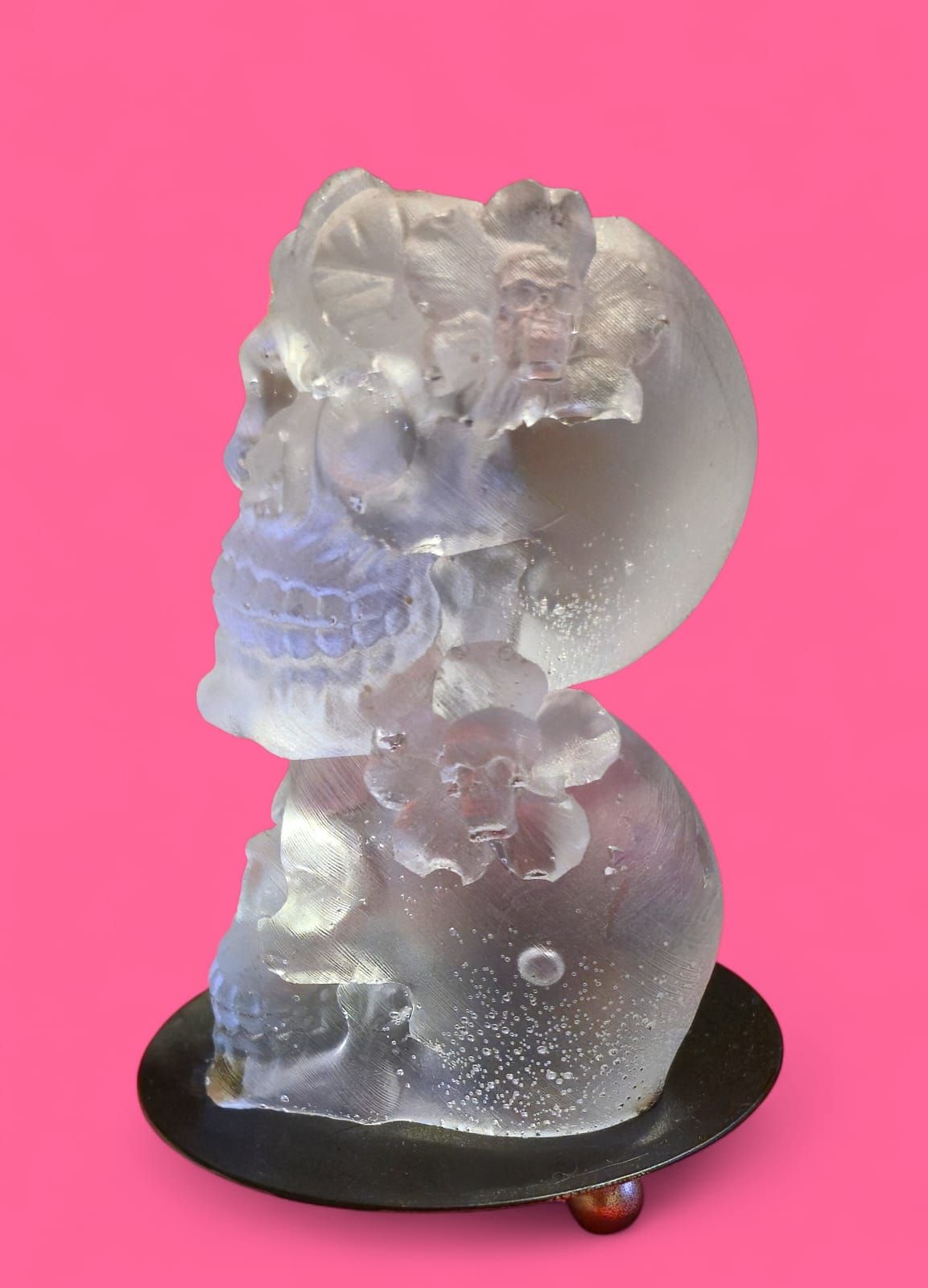 Dos portavelas de cristal transparente con forma de calavera sobre plato negro y fondo rosa.