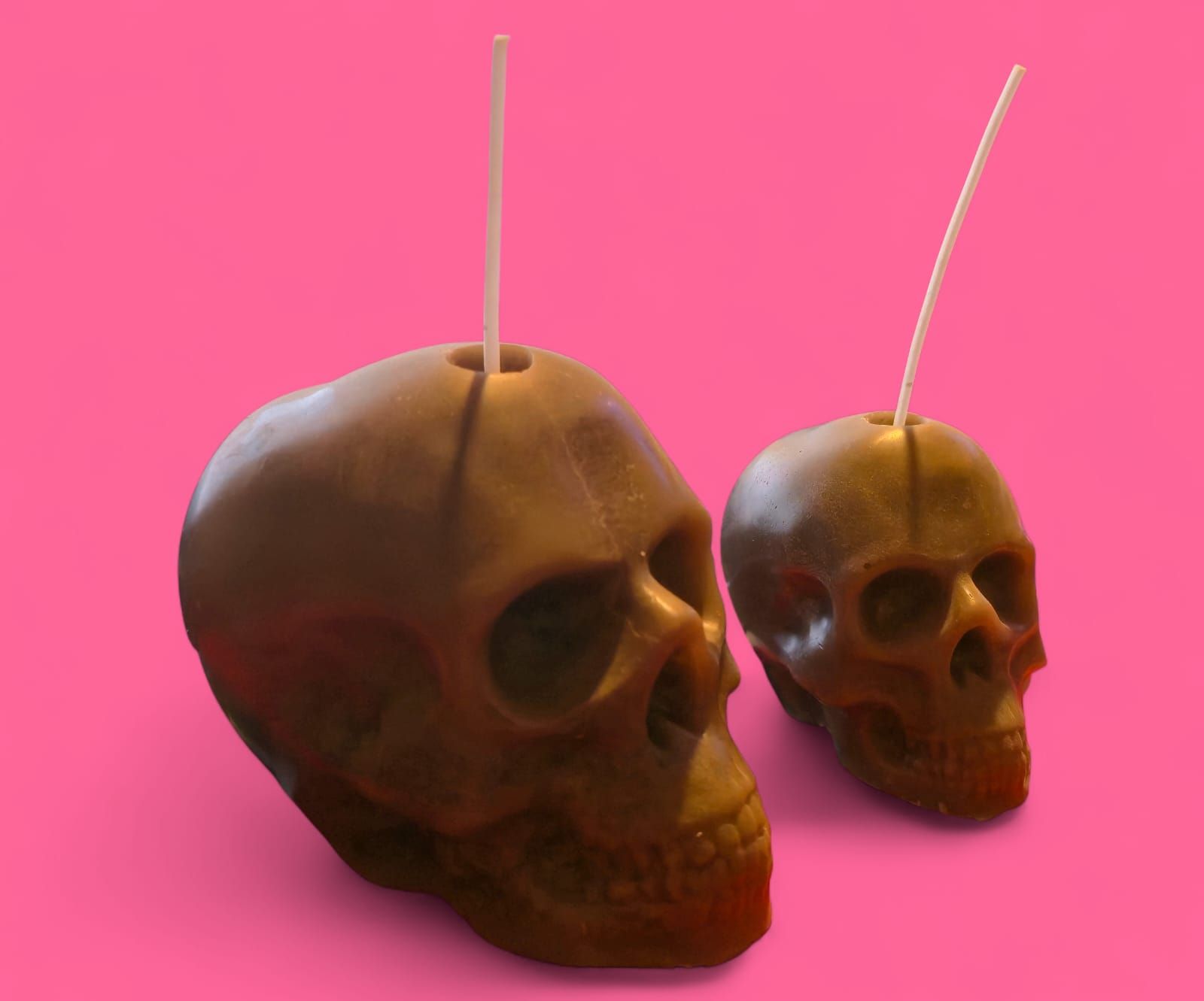 Dos velas marrones en forma de calavera con mechas blancas sobre fondo rosa.